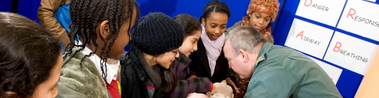 A member of London Ambulance Service showing a group of children performing first aid on a dummy