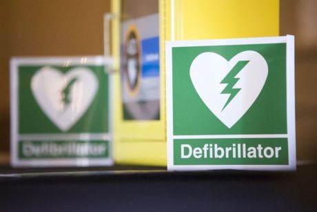 A defibrillator in a yellow cabinet with a green and white sticker identifying it as a defibrillator