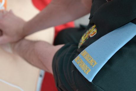 The shoulder epaulette of an Emergency Responder in the foreground as the person wearing the uniform is shown placing their hands on a chest to perform CPR 