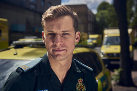 A close up shot of Cornelius in LAS uniform looking at the camera. Ambulance vehicles are shown behind him.