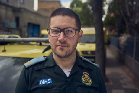 A close up shot of Ben in LAS uniform looking at the camera. Ambulance vehicles are shown behind him.