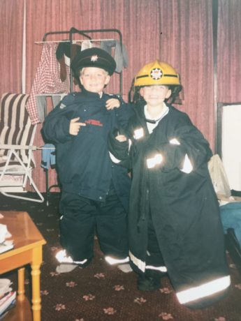 Tom and Jack as young children wearing oversized London Fire Brigade uniform including dark jackets and yellow firefighter hats