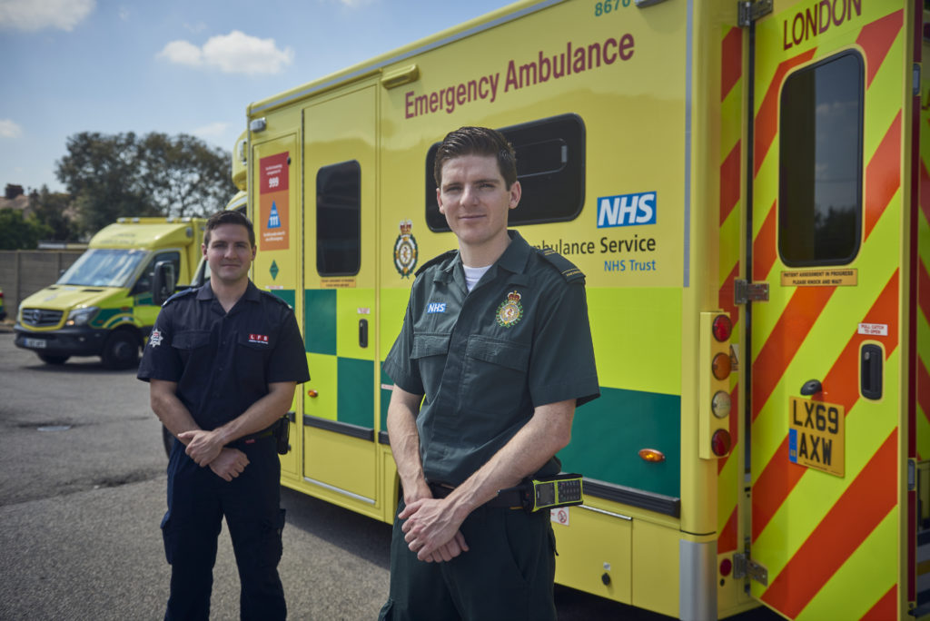 Tom (left) and Jack (right) stood in their uniforms in front of an ambulance. The ambulance is parked alongside them with its rear door open. 