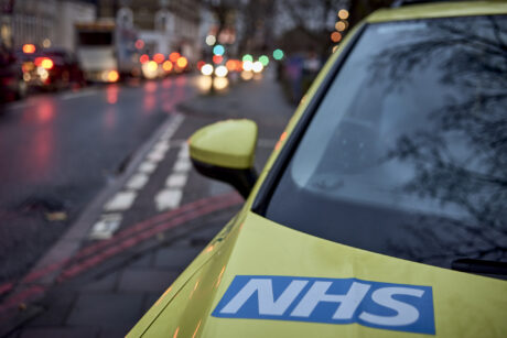 An ambulance car parked on a street with the NHS logo on the bonnet visible