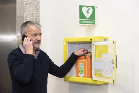 A man taking a defibrillator out of a cabinet attached to the wall while on the phone to the emergency services
