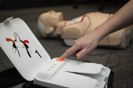 A person shown operating a defibrillator with a resuscitation dummy in the background