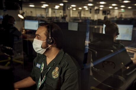 A call handler, wearing a mask and a headset photographed at his workstation with the rest of the control room in the background