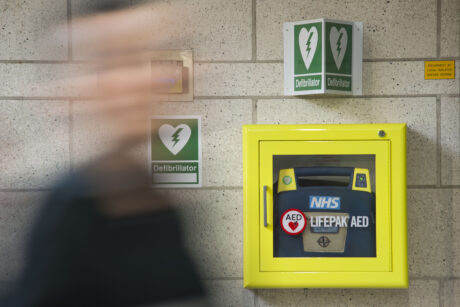 A defibrillator in a yellow cabinet attached to a wall as a person walks past