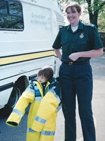 image showing a very young Millie in her mum's ambulance jacket stood next to her mum in front of ambulance