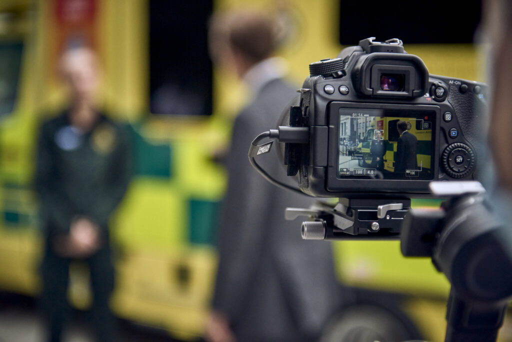 A photograph of a camera pointing towards two figures stood by an ambulance