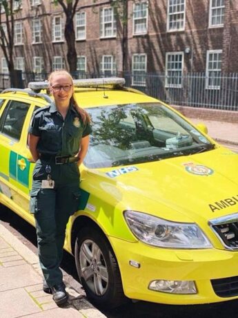 Paramedic Grace in her Service uniform posing for a photograph next to an ambulance car