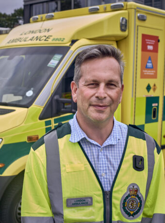 Daniel in a yellow LAS tabard smiles in front of a row of ambulances