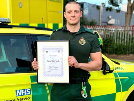 Gary Edwards in uniform stood in front of an ambulance car holding a certificate