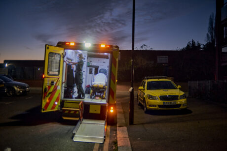 An ambulance and ambulance car parked next to each other in a dark street. The ambulance rear doors and lift are open and down and a paramedic is shown in the back