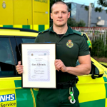 Gary Edwards in uniform stood in front of an ambulance car holding a certificate