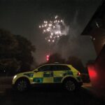 An ambulance car on a dark street with a firework going off in the sky behind