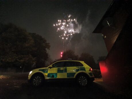 An ambulance car on a dark street with a firework going off in the sky behind