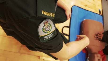 A paramedic (face not shown) seen crouched showing a person (face also not shown) how to do chest compressions on a dummy