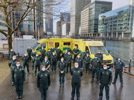 A group of AAPs in ambulance uniform stands in front of parked ambulances for a photograph in London's dockside