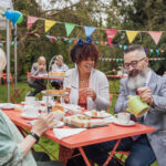 A young mixed race female and Caucasian hipster male sit chatting with a senior lady in the garden at a table filled with cakes and sandwiches, enjoying afternoon tea. The guy is pouring a cup of tea and there is colorful bunting hanging around the garden and people in the background sat eating also.