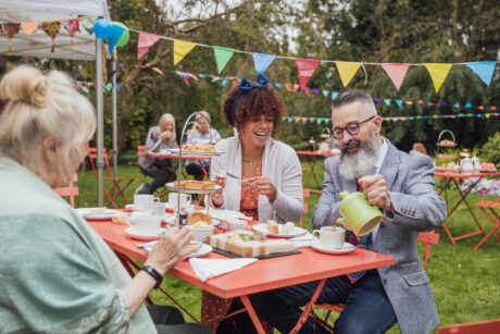 A young mixed race female and Caucasian hipster male sit chatting with a senior lady in the garden at a table filled with cakes and sandwiches, enjoying afternoon tea. The guy is pouring a cup of tea and there is colorful bunting hanging around the garden and people in the background sat eating also.