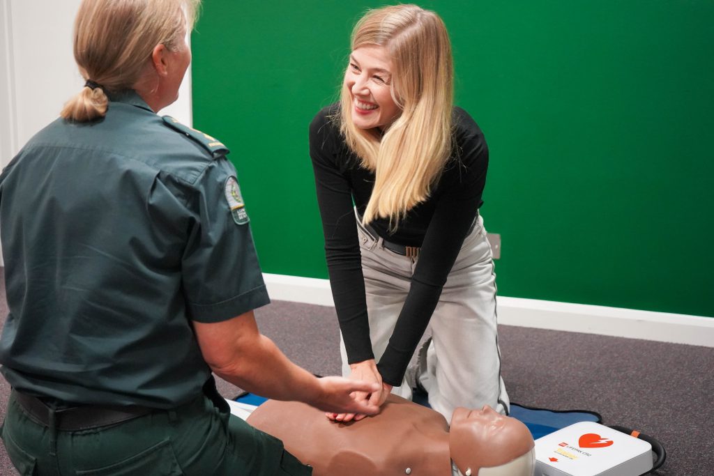 A picture of Rosamund Pike being taught lifesaving CPR skills by Sam Palfreyman-Jones. 