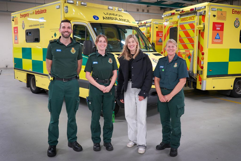 A picture of Rosamund Pike with Tom and Erica who she joined on an ambulance shift, and Sam who gave Rosamund London Lifesavers training. They are standing in front of a row of ambulances. 