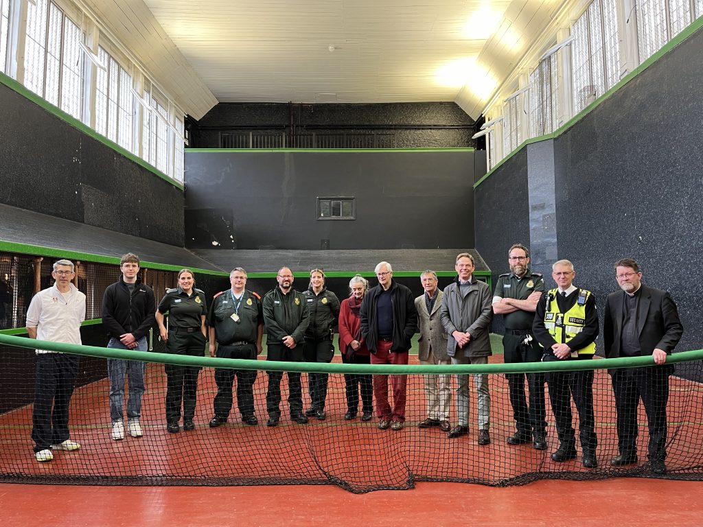 A group photo of cardiac arrest survivor David, London Ambulance Service staff, and Hampton Court Palace staff on a Real Tennis Court. 