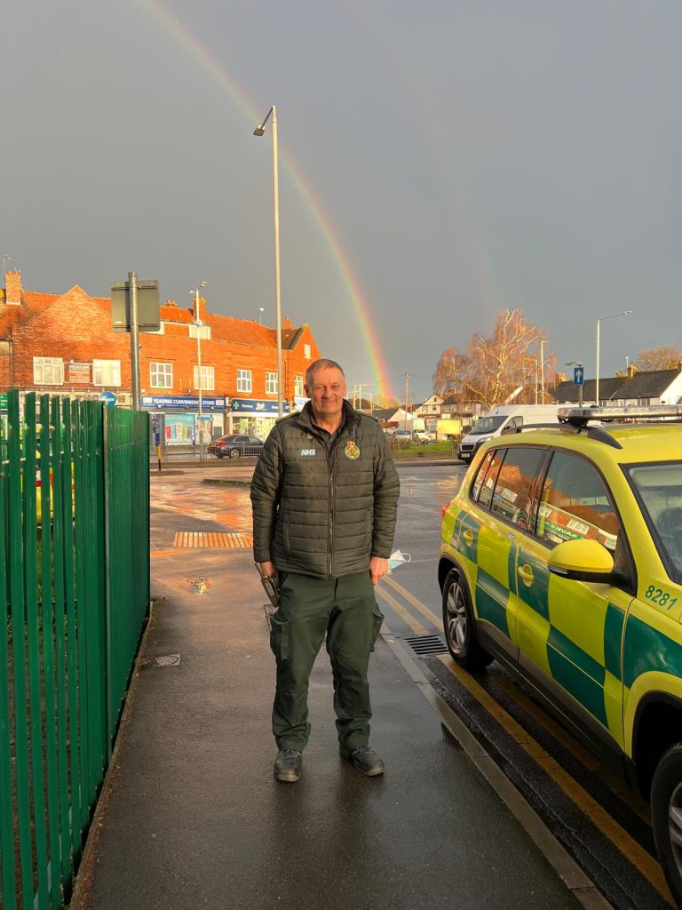 Paramedic Andy posing in front of a rainbow and next to a fast response ambulance